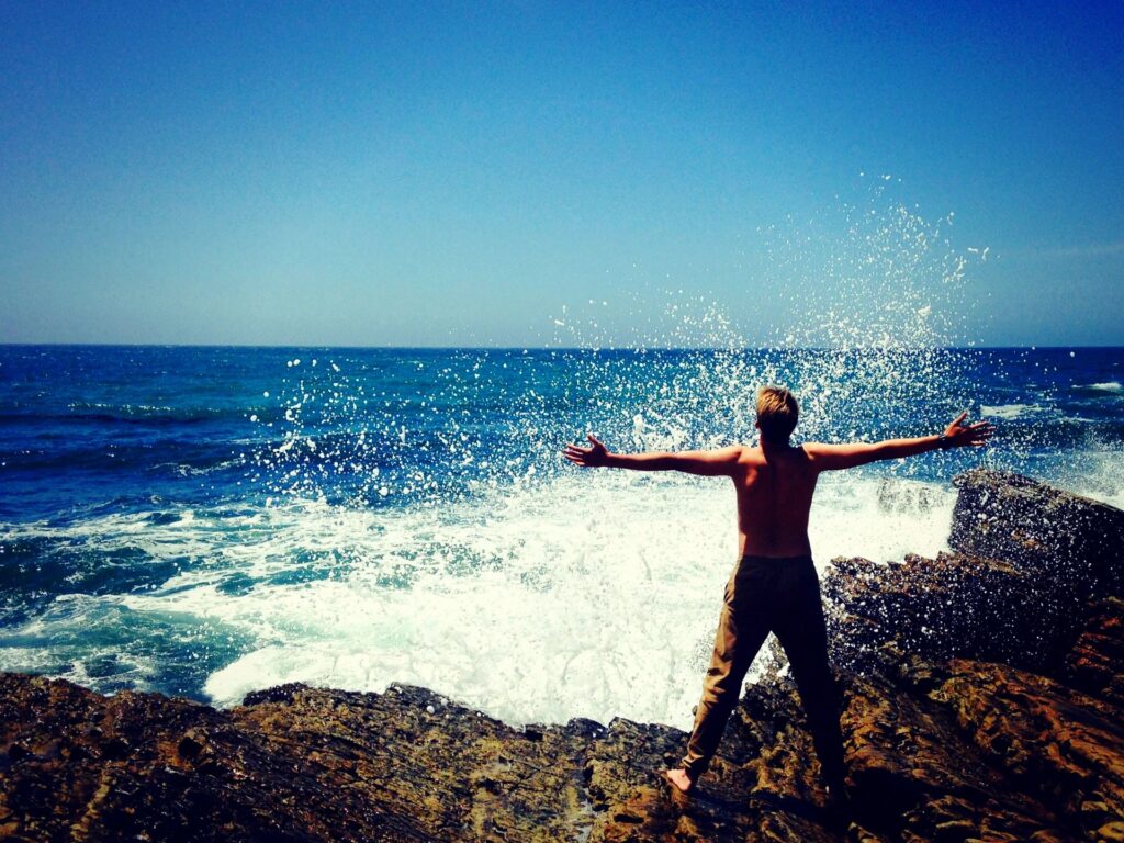 man standing proudly on rocks looking out to the ocean with arms outstretched.